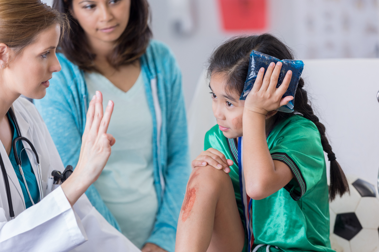 Young female soccer athlete is dazed while ER doctor asks her questions. The girl has an icepack on her head. The girl's mom is in the background.